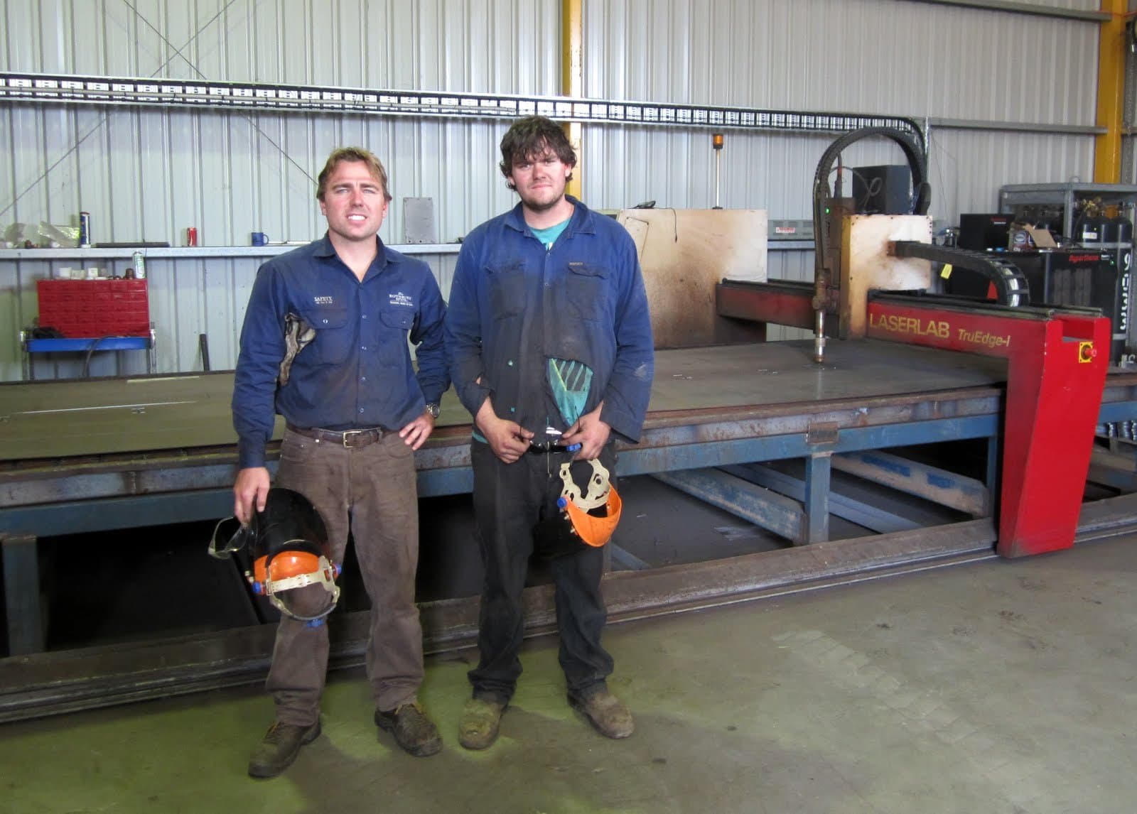 David and Rohan in work shirts, holding welding face shields, in front of a LaserLab TruEdge CNC plasma table